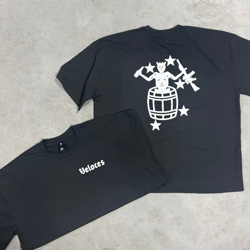 Two black t-shirts laid flat on a gray concrete surface. The larger shirt shows a white graphic of a skeleton playing a drum with rifles and stars on the back, while the smaller folded shirt displays the white wordmark 'Veloces' on the chest.